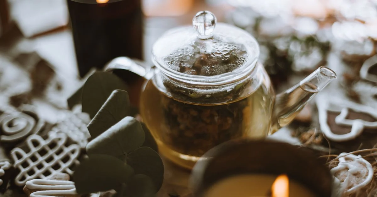 Une théière en verre entourée de bougies et de feuilles vertes sur une table rustique en bois.