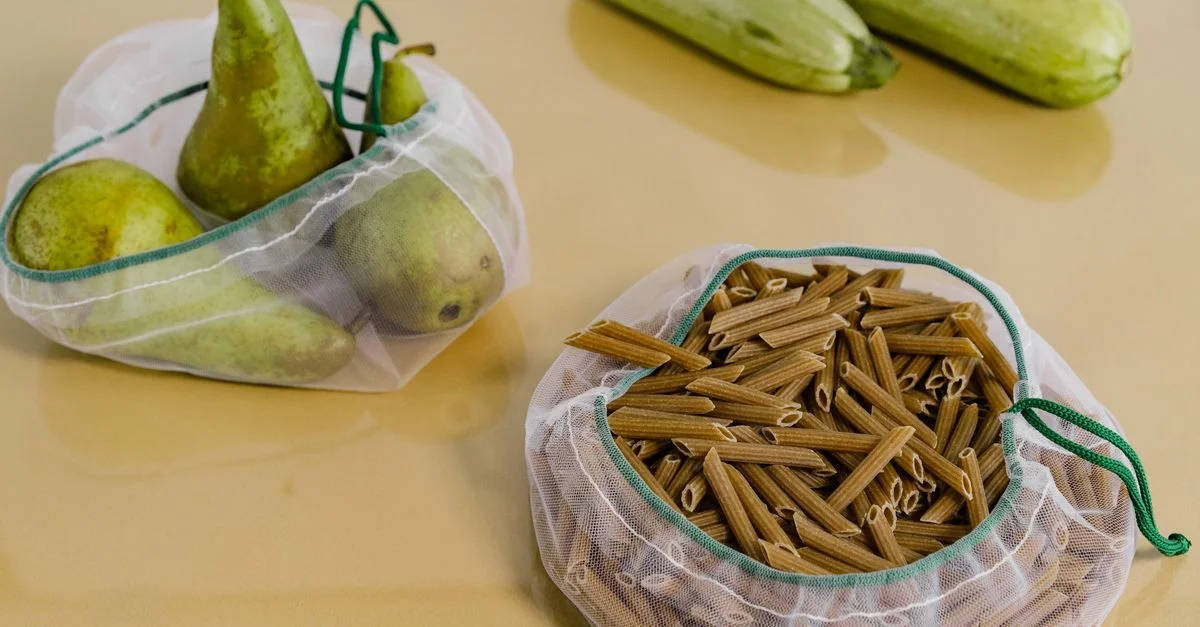 Sacs en filet réutilisables avec des poires, des pâtes penne et des courgettes sur un fond beige.